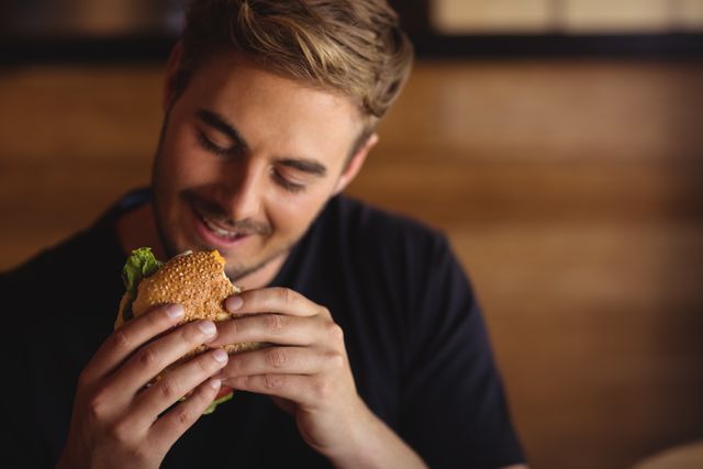 Man holding and looking at burger with a smile in a restaurant. Ideal for use in advertisements for restaurants, fast food chains, or lifestyle blogs focusing on dining and food enjoyment.