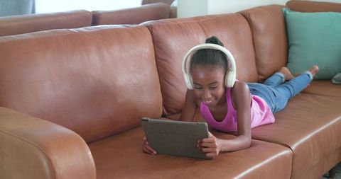 Child listening with headphones while using tablet on brown leather couch at home