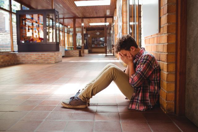 Schoolboy sitting alone in a school corridor, looking sad and stressed. Useful for illustrating themes of bullying, mental health issues, teenage struggles, and the emotional challenges faced by students. Can be used in educational materials, mental health awareness campaigns, and articles about youth and school life.