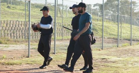 Adult baseball teammates walking to practice holding glove and bat on sunlit field