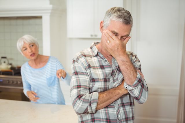 Senior couple having a heated argument in a modern kitchen. The man appears frustrated, covering his face with his hand, while the woman is gesturing and speaking emphatically. This image can be used to illustrate themes of relationship challenges, communication issues, and emotional stress in older adults. Suitable for articles on family dynamics, mental health, and elder relationships.