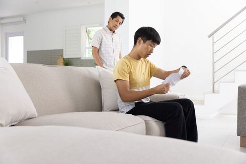 Two asian men in modern living room, young man reading letter while older man watching