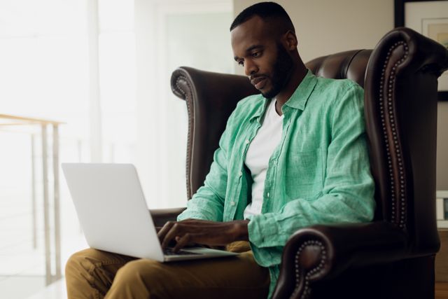 African-American man sitting on a leather chair using a laptop. Ideal for illustrating remote work, home office setups, modern technology use, and professional concentration. Suitable for business, technology, and lifestyle content.