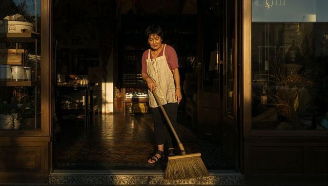 Senior woman sweeping shop threshold at golden hour with straw broom and apron rustic sunlit tiles