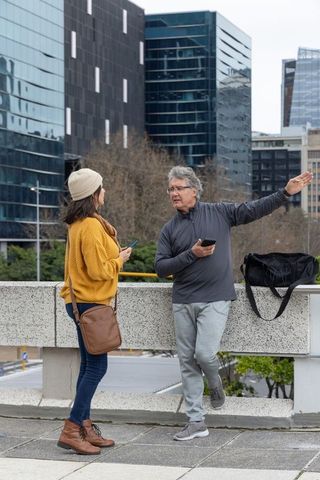 Senior couple conversing on urban balustrade while holding smartphones and duffel bag