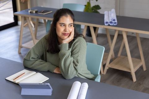 Designer sitting at studio desk in green sweater, looking pensive while planning blueprints