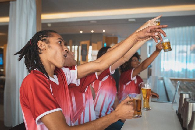 Diverse group of male and female sports fans raising glasses and watching a game at a bar. They are cheering and showing excitement, creating a lively and energetic atmosphere. This image can be used for promoting sports events, bar and restaurant advertisements, social gatherings, and community events.