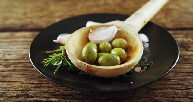 Green olives and garlic arranged in rustic wooden spoon on black plate with herbs in background. Suitable for use in culinary blogs, food magazines, Mediterranean recipes, promotional material for organic food products, or social media content related to cooking and food styling.
