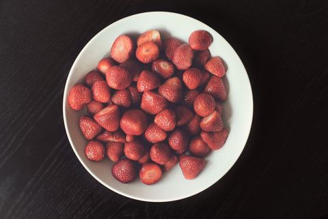 White bowl holding ripe red strawberries on dark wooden table, overhead food styling