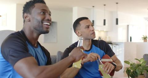 Diverse friends cheering while watching football in open-plan lounge wearing blue jerseys