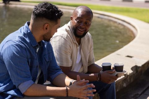Two diverse men sitting by fountain sharing coffee and laughing in casual conversation
