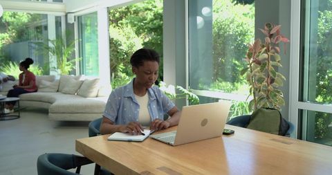 Black young woman working on laptop and writing in notebook in modern home workspace