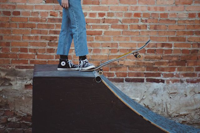 Teenager wearing denim jeans and black sneakers standing on a skateboard, preparing to perform a trick on a skate ramp. Background includes a brick wall, suggesting a gritty urban environment. Useful for promoting outdoor activities, youth culture, and skateboarding communities or clothing brands targeting teenagers.
