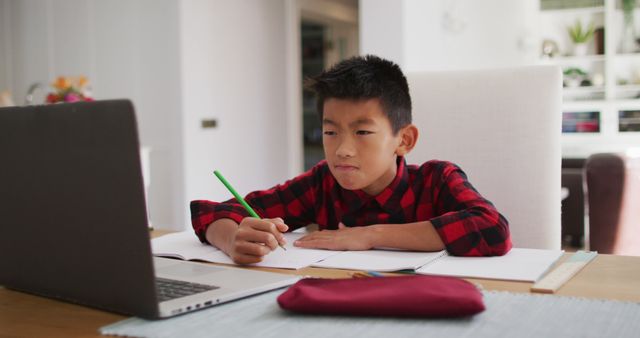 A young boy in a red plaid shirt is diligently studying and taking notes while attending online classes at home. This can be used to depict home learning, remote education, or virtual schooling. It is suitable for educational content, articles on homeschooling, e-learning platforms, and resources for parents managing children's education from home.