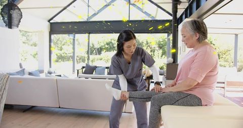 Homecare caregiver assisting senior woman with leg mobility exercise at home in bright living room