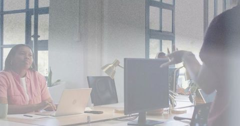 Woman typing while colleague gesturing in modern open office with laptop and monitor
