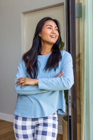 Smiling asian woman standing at sliding door in light blue loungewear with plaid pants