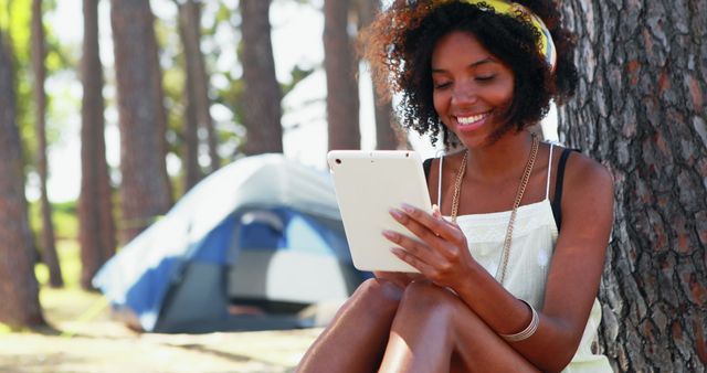 This image depicts a woman sitting against a tree while using a digital tablet in a forest camping scene. Perfect for websites and articles focused on technology, outdoor activities, camping, relaxation, natural lifestyle, and leisure time.