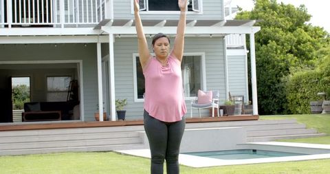 Mature woman stretching arms overhead on lawn by modern house and plunge pool