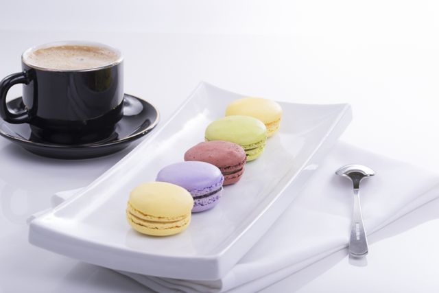 Image featuring a variety of colorful macarons arranged in a line on a rectangular white plate, beside a cup of coffee. A spoon is placed next to the plate, enhancing the elegant presentation. Perfect for use in food blogs, coffee shop promotions, and gourmet dessert advertisements.