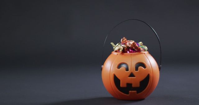 This depicts a festive orange pumpkin bucket filled with an assortment of colorful candies. The smiling jack-o'-lantern face reflecting the Halloween spirit can be used in promotional materials for Halloween events, decorating tips, or party invites. Perfect for illustrating articles or blogs discussing Halloween traditions, trick-or-treat safety, or candy consumption during October festivities.