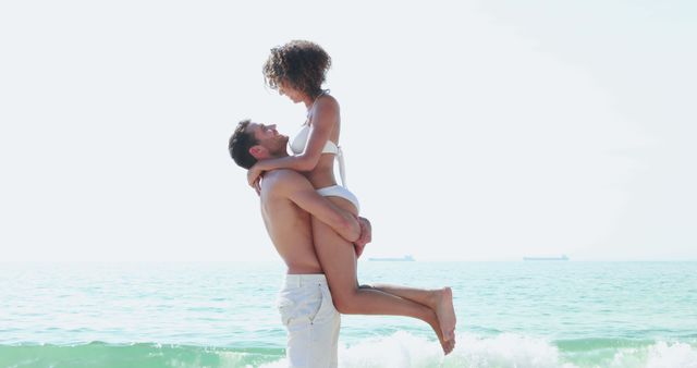 A young Caucasian couple enjoys a romantic moment on a sunny beach, with the man lifting the woman in his arms, with copy space. Their joyful expressions and the serene ocean backdrop evoke a sense of love and happiness.