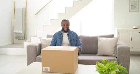 African man smiling while opening delivery box on sofa in bright modern living room