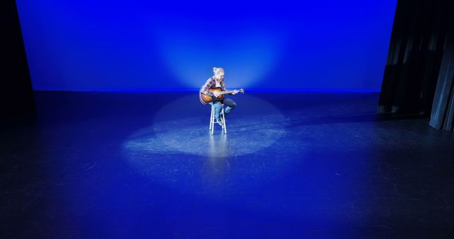 A musician sits center stage under a spotlight, playing the guitar in a serene setting, with copy space. The image captures a moment of artistic expression, highlighting the performer's solitary presence in the vastness of the stage.