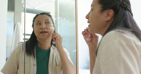 Mature woman using rose quartz facial roller at bathroom vanity mirror for skincare ritual