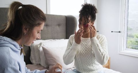 Female friends applying skincare and under-eye patches while sitting on cozy bed at home