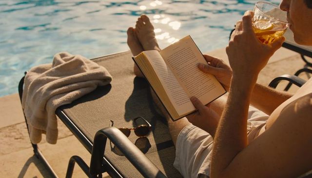 Sunlit poolside reading: shirtless man relaxing with book and drink at golden hour