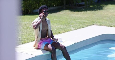 Young man sitting poolside talking on phone and holding drink, sunglasses and swim trunks