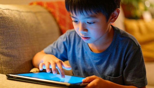 Asian boy using tablet on couch at night showing blue screen glow and concentration