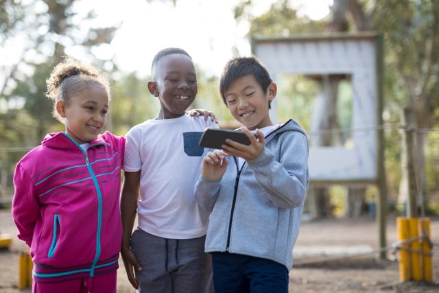 Three children are standing together in a park, smiling and looking at a mobile phone. They are dressed in casual clothing and appear to be enjoying their time outdoors. This image can be used for themes related to childhood, technology, friendship, and outdoor activities. It is ideal for educational materials, advertisements for children's products, or articles about the impact of technology on young people.