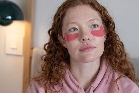 Relaxed redhead applying pink under-eye gel patches during cozy at-home skincare ritual