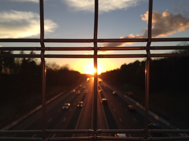The image captures the sun setting over a busy highway with vehicles traveling in both directions, viewed through a wire fence. This could be used in travel blogs, articles on daily commutes, urban planning, or transportation services.
