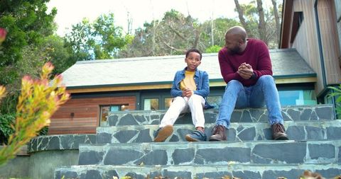 African american father and son sitting on stone steps outside modern wooden home bonding