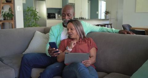 Diverse couple relaxing on sofa using smartphone and tablet in modern living room