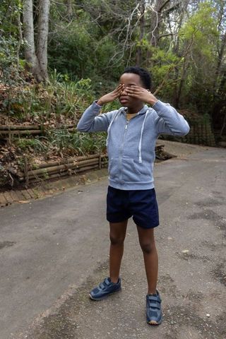 African american boy covering eyes on park path wearing hoodie, shorts and sneakers