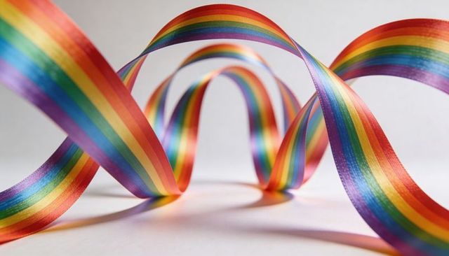 Looping rainbow satin ribbons twisting and arching over white cardstock with soft shadows