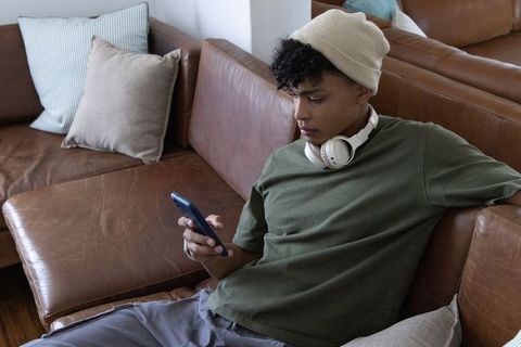 African american man lounging on leather sofa wearing beanie and headphones using phone