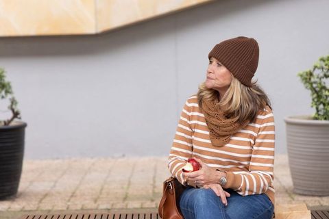 Senior woman sitting on urban bench holding half-eaten apple wearing knit hat and scarf