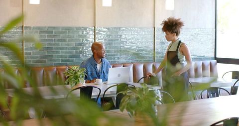 Barista talking to remote worker at cafe booth with laptop and plants, modern coworking