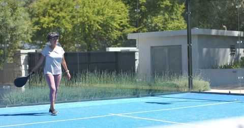 Walking woman carrying black paddle on bright blue paddle tennis court in sunny backyard