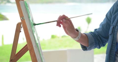 Mature woman painting green landscape on canvas at easel by lakeside in denim shirt