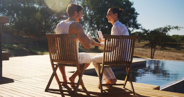 A young Caucasian couple enjoys a conversation while sitting on wooden chairs by a poolside, with copy space. Their relaxed posture and the serene outdoor setting suggest a moment of leisure and connection.
