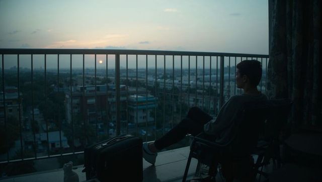Teen on highrise balcony watching sunset with suitcase, contemplative urban travel moment