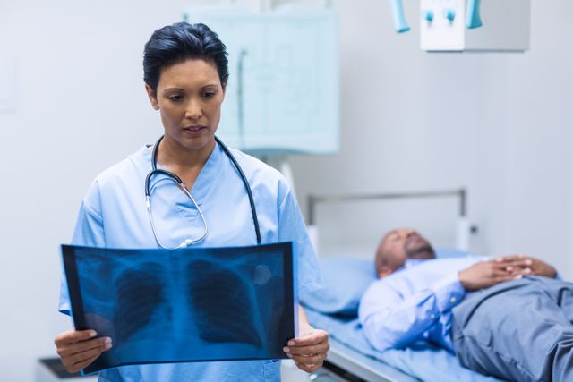 Female doctor examining chest x-ray in hospital with patient lying on bed in background. Ideal for use in healthcare, medical, and hospital-related content, showcasing medical professionals, patient care, and diagnostic procedures.