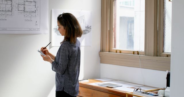 Female architect stands beside desk reviewing blueprints in open, well-lit office. Using clipboard for efficient planning. Ideal for concepts related to profession, design, creative work, and modern office environment.