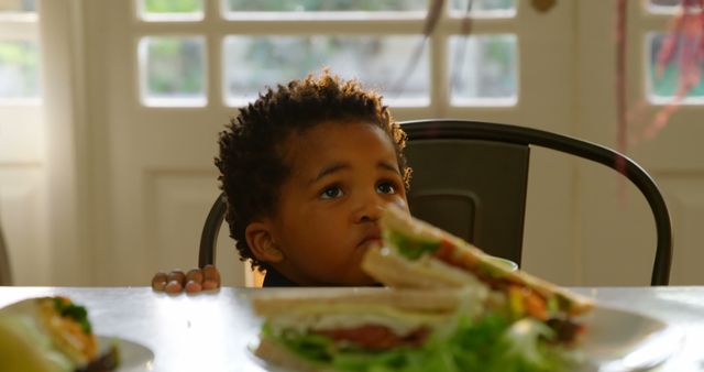 Front view of cute little black boy sitting on chair at dining table in a comfortable home. Food plate on dining table 4k